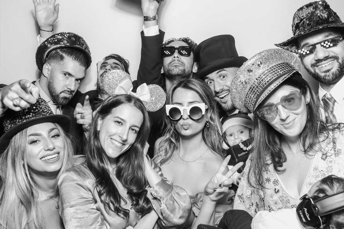 Black-and-white photo booth image of a lively wedding group in Mississauga wearing fun props like glitter hats, sunglasses, and novelty accessories, including a baby in the front row.