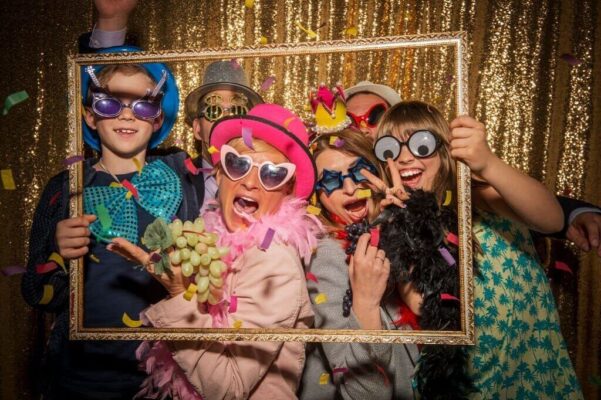 Group of friends and kids wearing playful costumes and sunglasses posing with props and confetti in front of a gold backdrop at a Kitchener birthday party photo booth