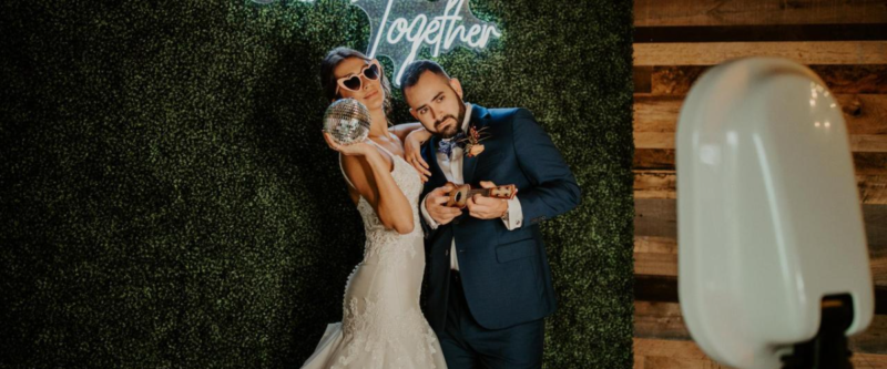 Bride holding a disco ball and groom with a mini guitar posing in front of a green wall with neon sign at their Markham wedding photo booth setup